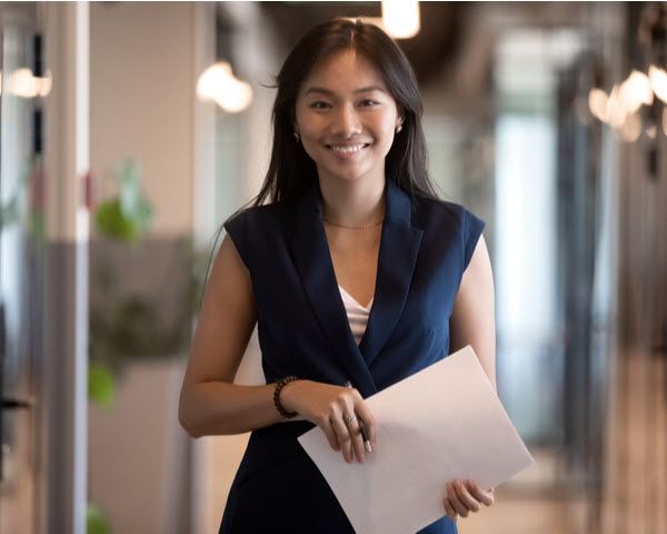 businesswoman looking at camera holding papers while standing in office hallway